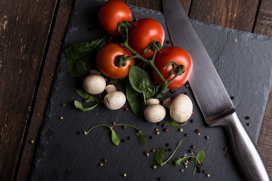 Top view of fresh tomatoes, mushrooms, and greens on a slate board with knife.
